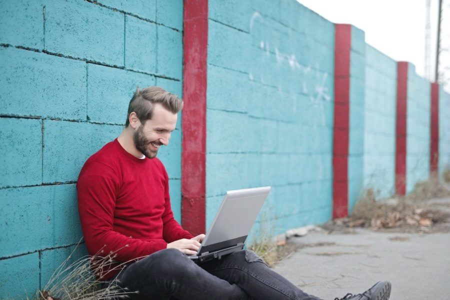 Man sitting against a blue brick wall typing on a laptop