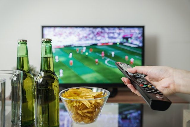 Person's hands with snacks holding a remote control in front of a TV