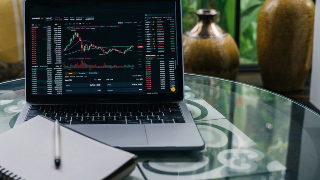 Laptop computer showing crypto data on its screen. A notebook and pen are to the left.