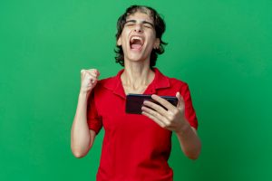 Joyful young caucasian girl with pixie haircut holding mobile phone clenching fist isolated on green background with copy space