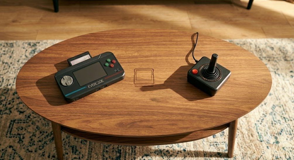 A high-angle, top-down photograph in a sunlit living room focusing on a mid-century modern coffee table where a classic black Atari-style joystick controller is positioned. A second, identical controller and a modern-looking Evercade VS console sit nearby, all ready for classic gaming on the large TV positioned to the side.