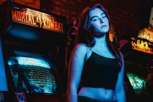 A young woman stands in a neon-lit arcade surrounded by retro gaming machines.